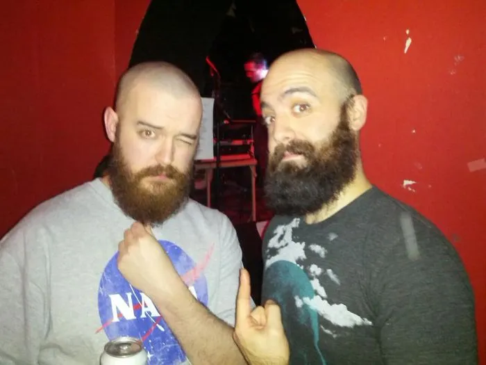 Two bearded strangers amazed at their real-life doppelganger encounter, sharing a fun moment indoors.