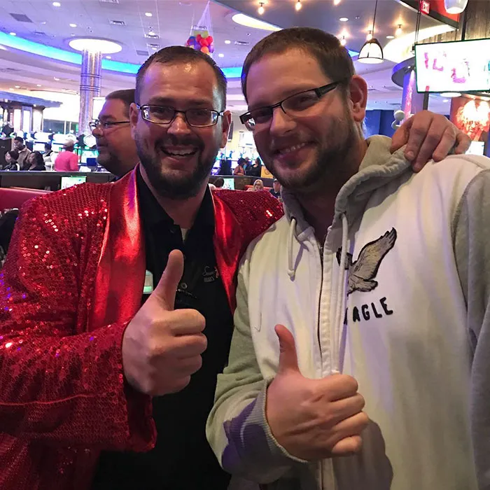 Two men, resembling doppelgangers, pose with thumbs up in a lively setting.