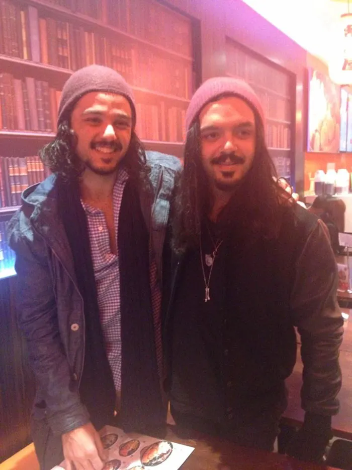 Two men in matching outfits, appearing as real-life doppelgangers, standing in a cozy library-themed setting.