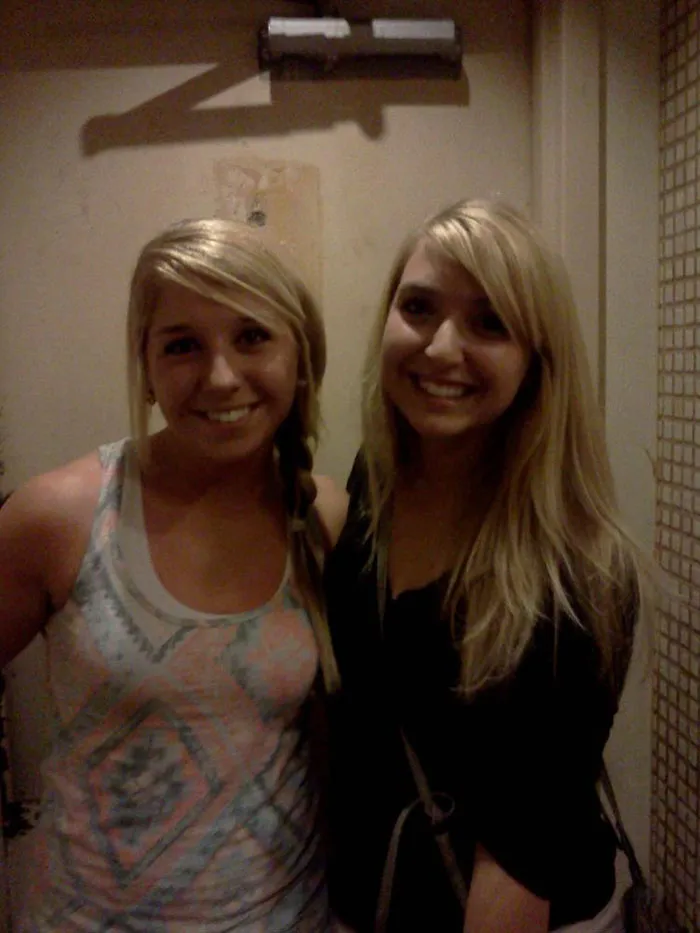 Two women standing together, resembling doppelgangers with similar hairstyles, smiling at the camera indoors.
