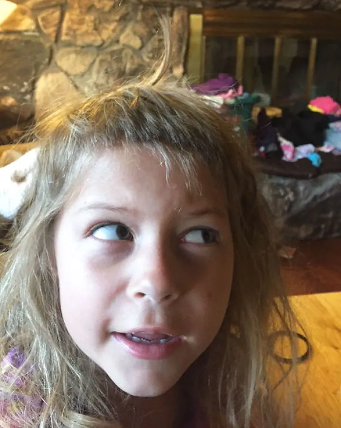 Young child with uneven self-cut bangs, looking sideways in a living room.