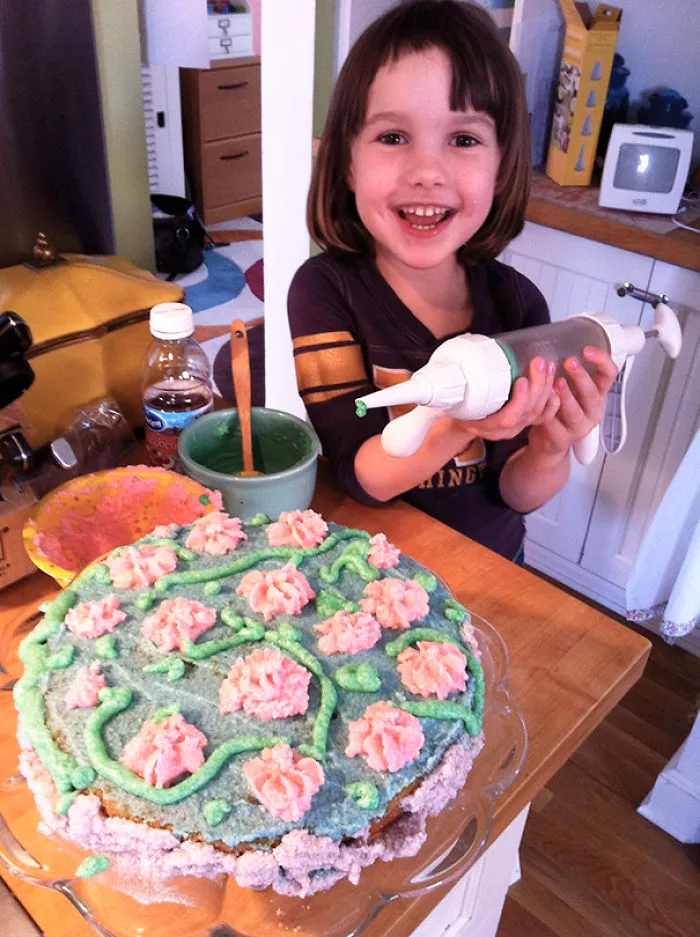 Child with uneven haircut holding a cake decorating tool, smiling next to a decorated cake.