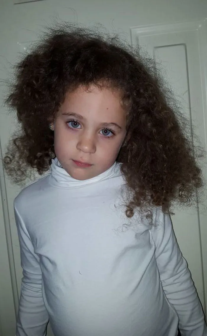 Child with uneven haircut, wearing a white turtleneck, looks regretful.