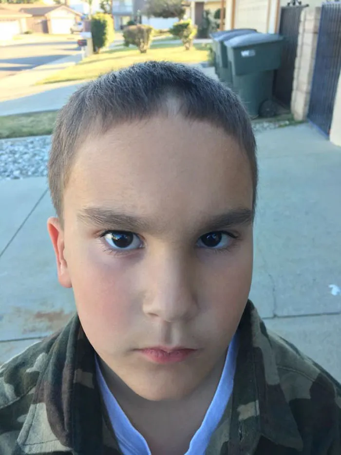 Child with a self-cut haircut, looking solemn in a camouflage jacket outdoors.