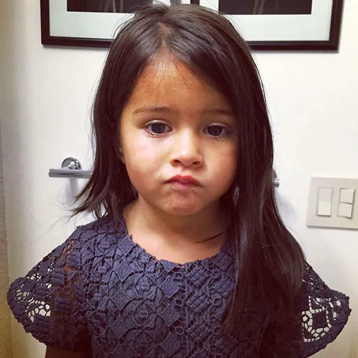 Child with uneven haircut, looking regretful, standing in front of a bathroom wall after cutting their own hair.