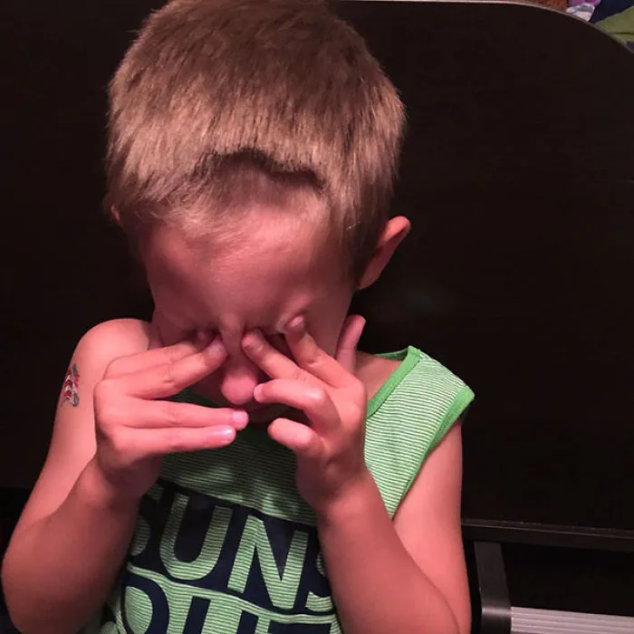 Child covering eyes after haircut mishap, wearing a green-striped tank top.