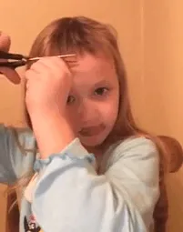 A young child cutting their own hair with scissors, appearing focused, in a casual home setting.