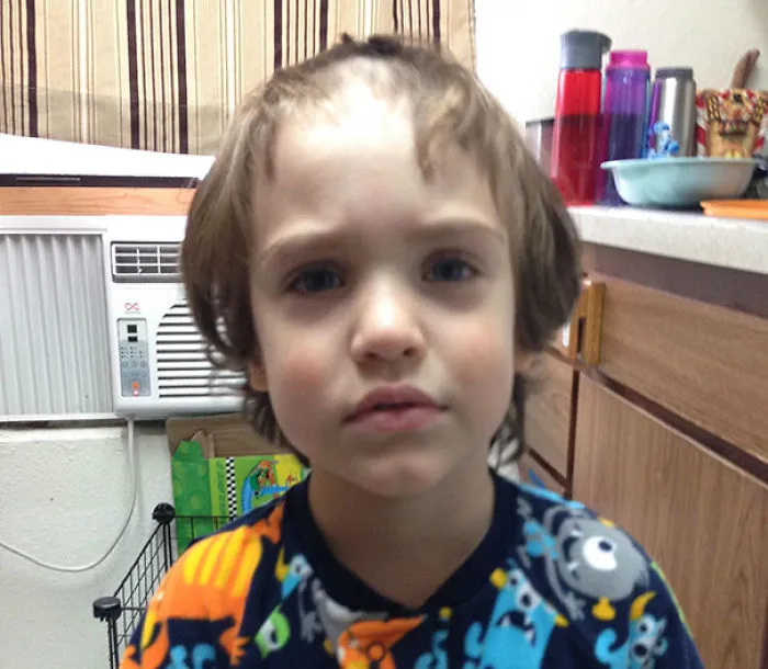 Child with uneven self-cut hair, wearing colorful pajamas, looking slightly regretful in a kitchen setting.