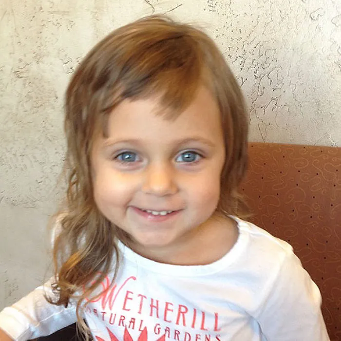 Young child with uneven self-cut hair, smiling in a white shirt.