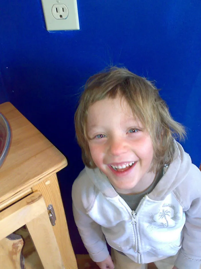 Child smiling with uneven self-cut hair, wearing a hoodie, standing near a wooden table against a blue wall.