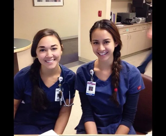 Two women in blue uniforms, smiling and sitting together, resembling real-life doppelgangers in a medical setting.