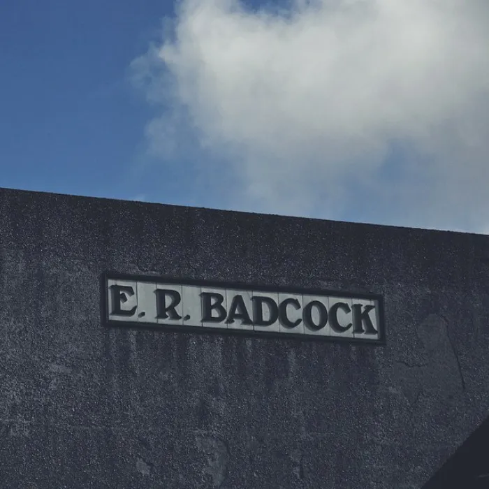 Sign displaying a humorous name, E.R. Badcock, against a cloudy sky.