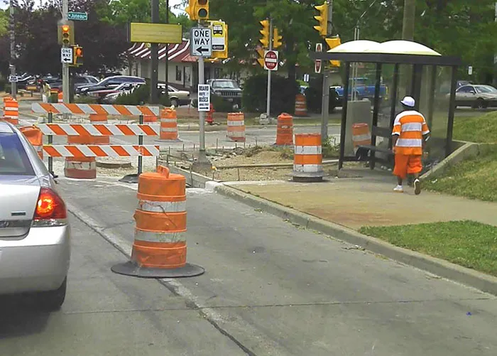 Man wearing orange stripes at a construction site, blending humorously with road barrels for a "who wore it better" moment.