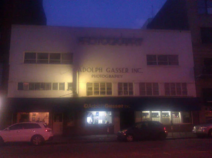Storefront at night with a sign displaying a funny name, featuring parked cars on the street.