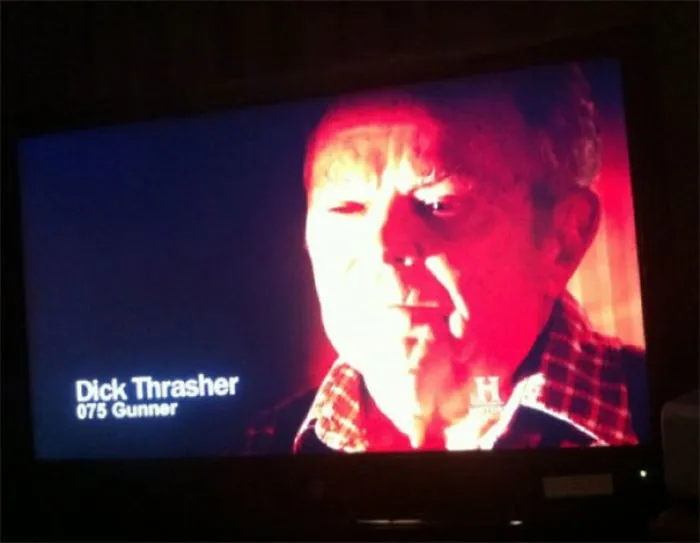 Man with funny name on TV screen labeled "Dick Thrasher," wearing a checkered shirt under red lighting.
