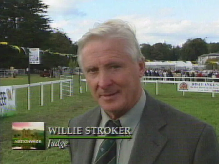 Man in a suit with a humorous name tag "Willie Stroker," standing outdoors at an event.