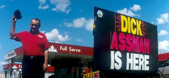 Man in front of a gas station sign with "Dick Assman is here," showcasing funny names.