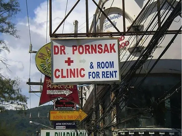 Funny name sign: "Dr. Pornsak Clinic & Room for Rent" on a busy street with other colorful signs.