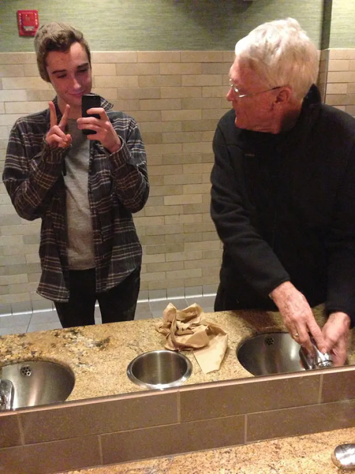 Boy making mirror selfie while old man washing hands 