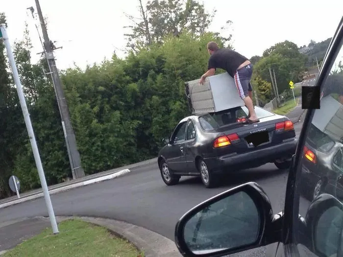 Man standing on a moving car roof holding furniture, illustrating risky behavior.