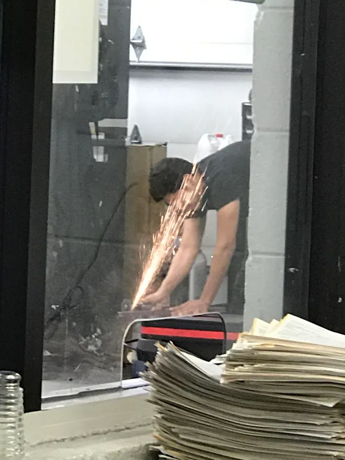 A person using a grinder, sparks flying in a workshop. Stack of papers in the foreground.