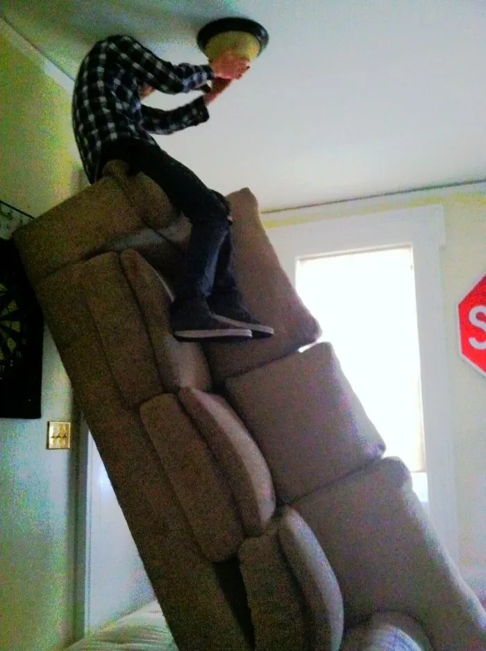 Man standing on stacked couches changing ceiling light, highlighting why women live longer.