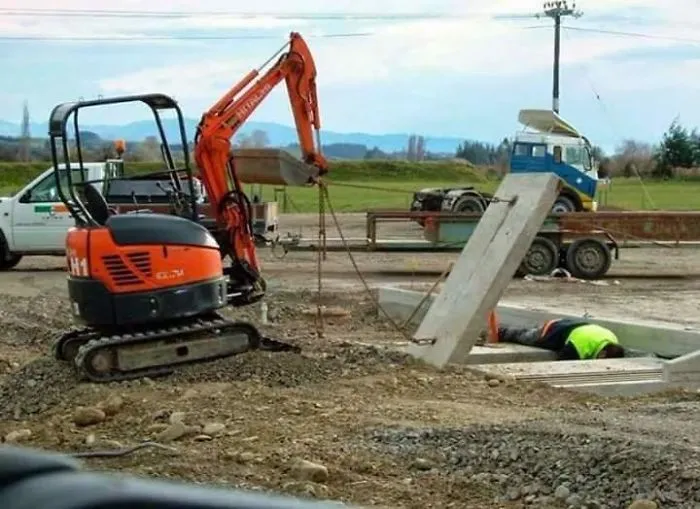 Excavator lifting concrete slab while worker in neon vest lies in ditch, illustrating reasons women may live longer than men.