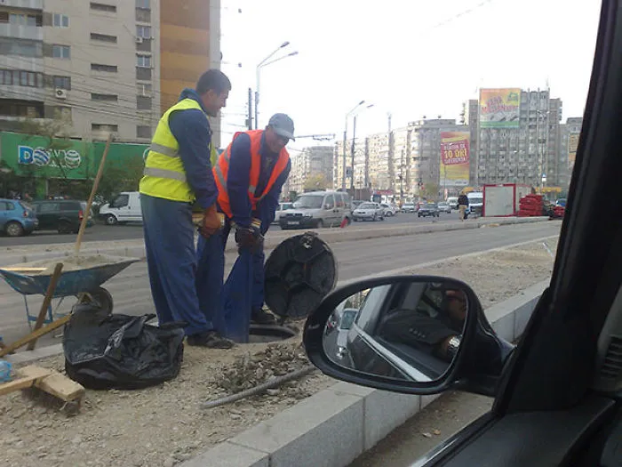 Two men in safety vests working on a roadside construction site, lifting a heavy object near a busy street.