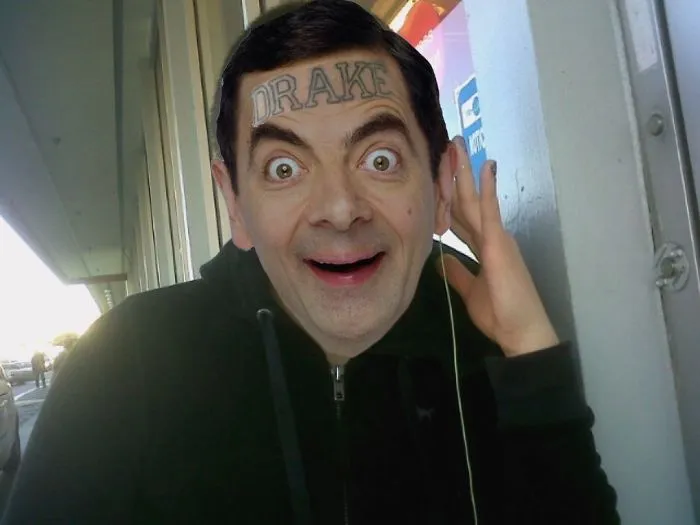 Mr. Bean photoshopped with a Drake tattoo on his forehead, smiling and wearing a black hoodie outdoors.