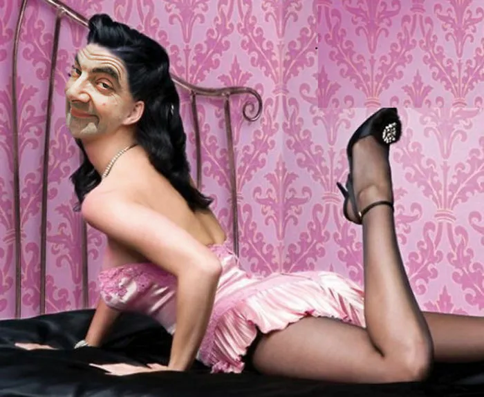 Photoshopped Mr. Bean posing in a pink dress with black stockings against a pink patterned wallpaper background.