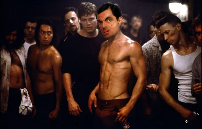 Mr. Bean photoshopped shirtless into a fight scene, surrounded by intense male characters in a dimly lit room.