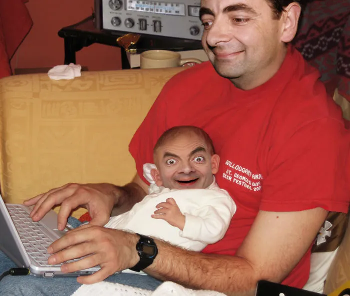 Man in red shirt using laptop holding baby with photoshopped Mr. Bean face, showcasing hilarious Mr. Bean photoshop edits.