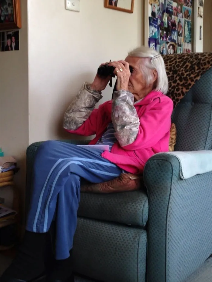 Older woman using binoculars indoors, embodying technology fail humor.