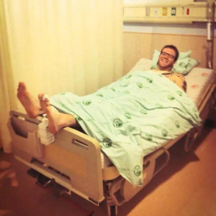 Tall man lying in a hospital bed with feet hanging over the edge, illustrating everyday struggles of tall people.