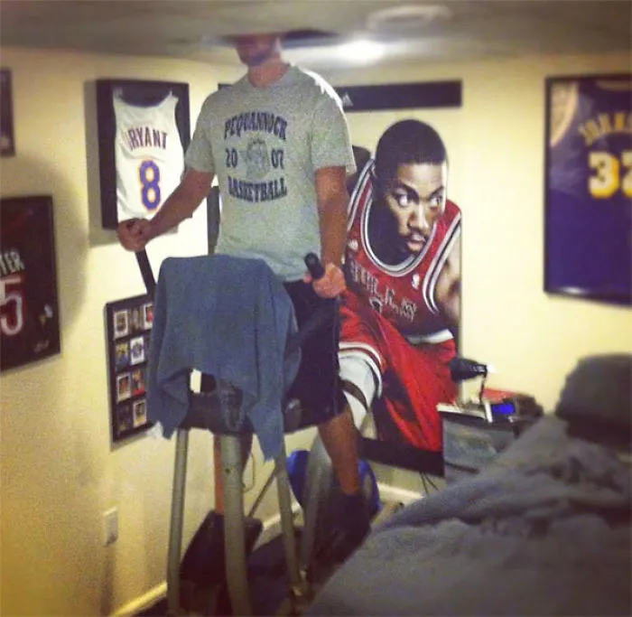 Tall person using an elliptical machine in a low-ceilinged room decorated with basketball jerseys and posters.