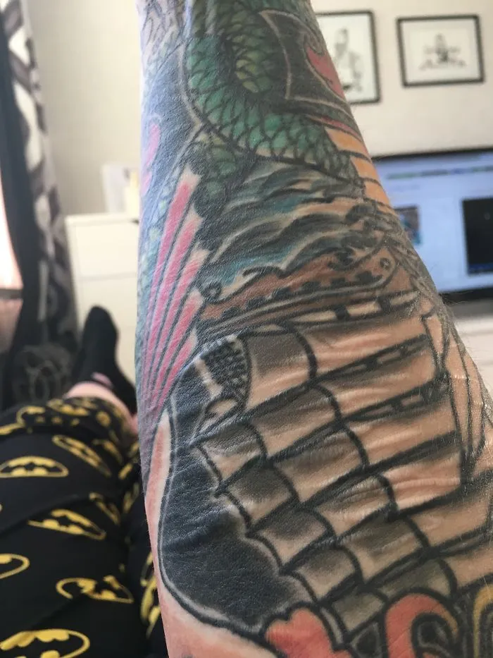 Tattoo art on arm transforming scar into a colorful design, with Batman-themed pants visible in the background.