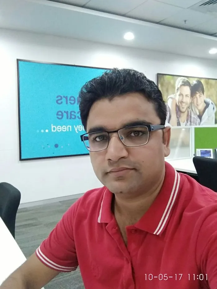 Man in office wearing glasses and a red polo shirt, sitting at a table with promotional posters in the background.