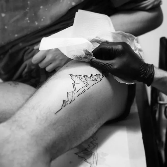 Artist creating a single continuous line tattoo on a person's leg with a black glove.
