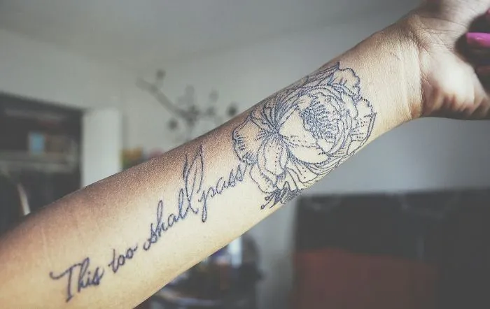 Tattoo art transforming a scar on an arm, featuring floral design and text "This too shall pass."