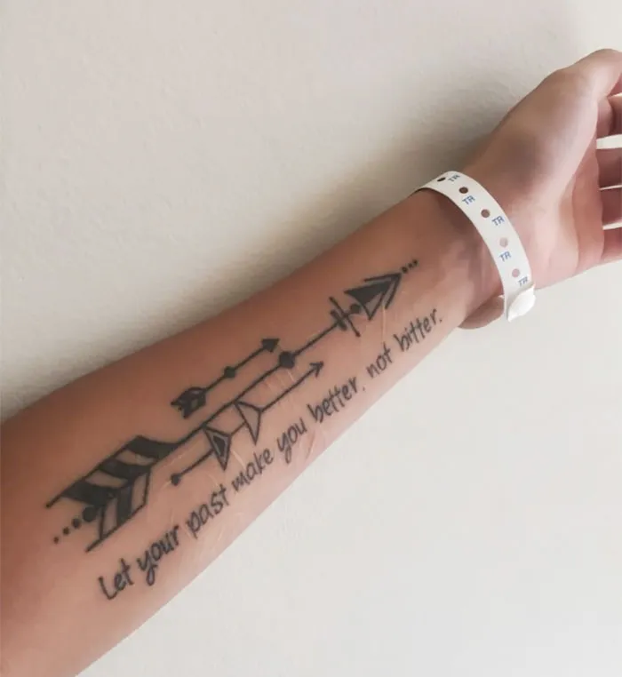 Tattoo on forearm transforms a scar with an arrow design and inspiring quote.