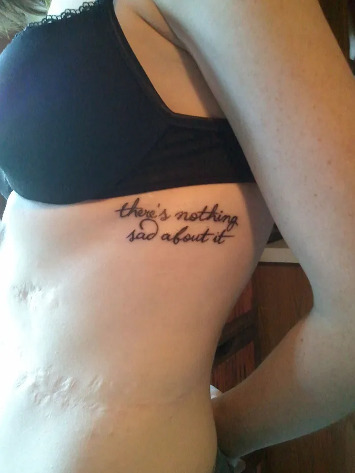 Tattoo on side with text "there's nothing sad about it" creatively incorporating and enhancing a scar.