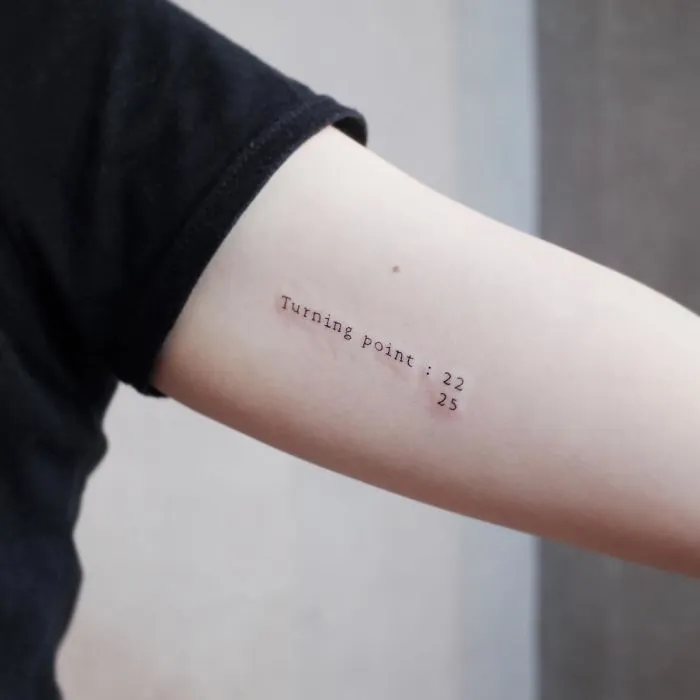 Minimalist tattoo on arm reading "Turning point: 22 25" in black ink.
