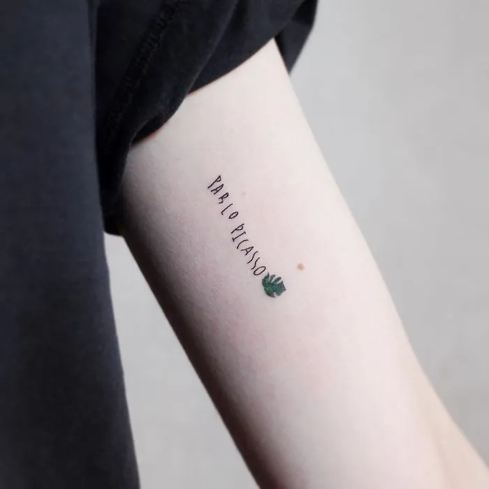 Minimalism tattoo with "Pablo Picasso" text on an arm featuring a small green leaf design.