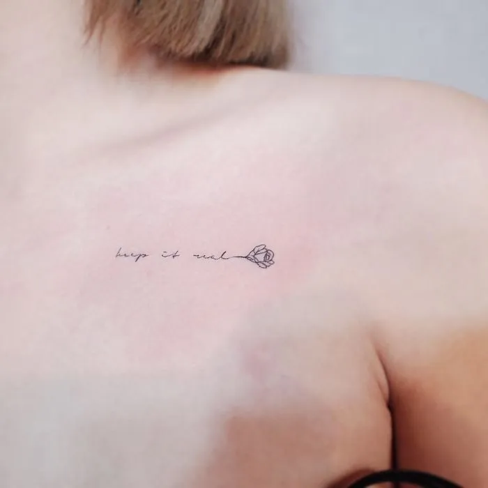 Minimalist tattoo on shoulder, featuring delicate script "keep it real" with a small flower, embodying tiny discreet design.