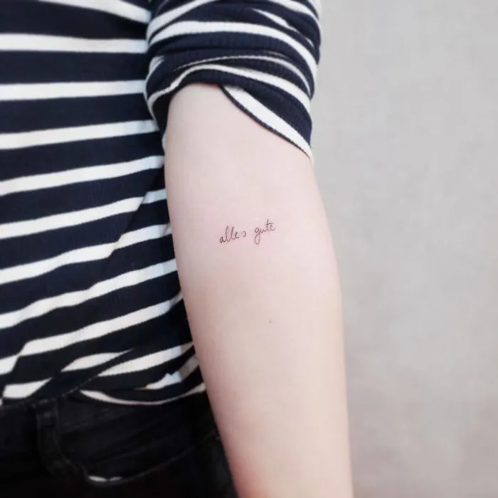 Minimalist tattoo on arm with "alles gute" in cursive, ideal for tiny discreet tattoo lovers.