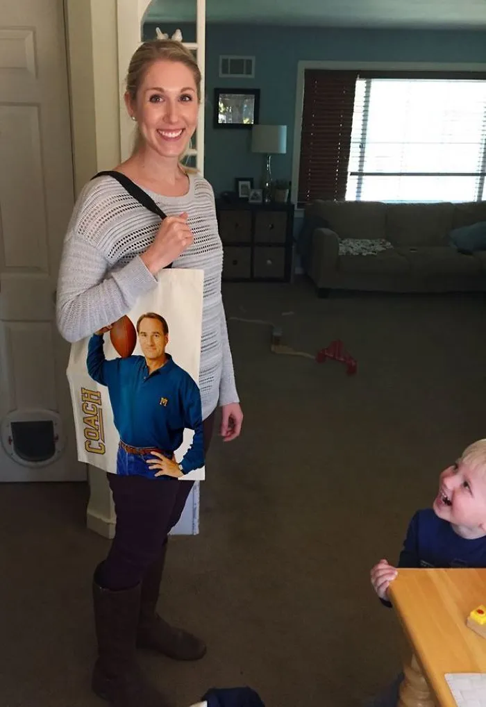 A woman smiling, holding a funny tote, while a child laughs nearby in a living room.