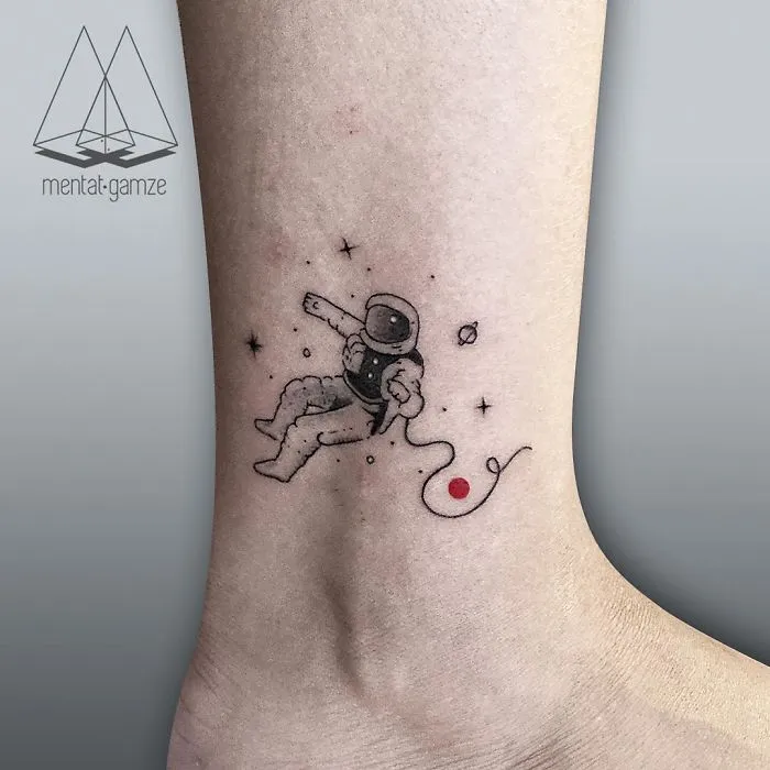 Astronaut tattoo with a red dot, surrounded by stars and planets, on the ankle.