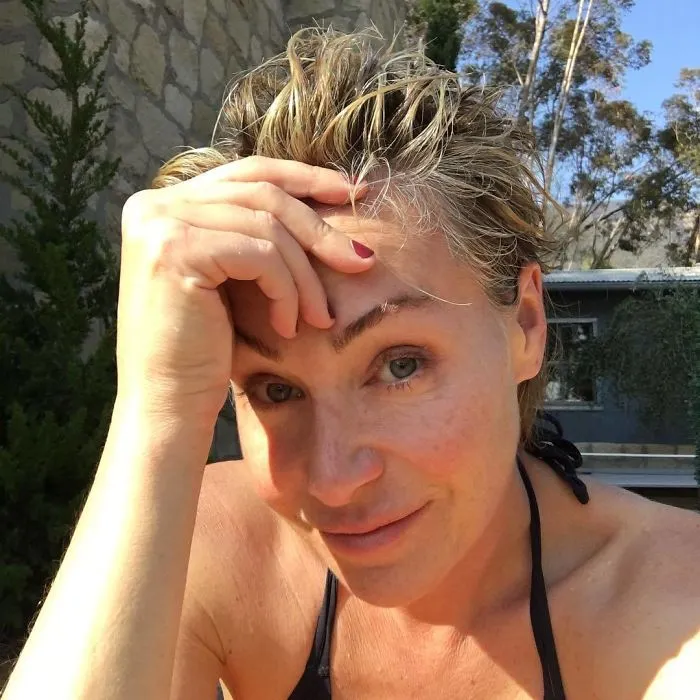 Celebrity without makeup, smiling outdoors with hand on forehead, wearing a black top.