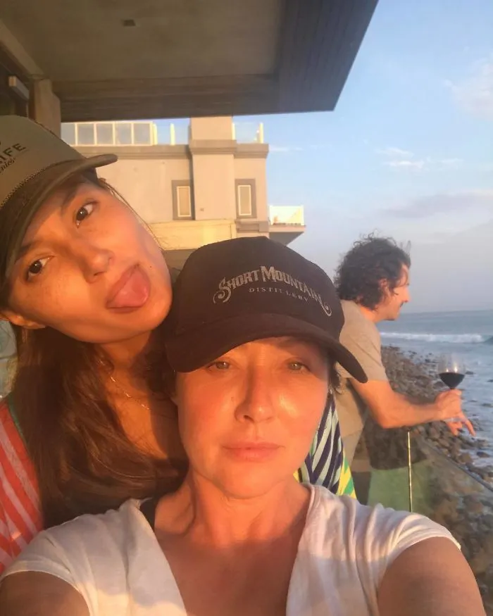 Two people by the ocean, wearing caps, without makeup, enjoying the view at sunset.