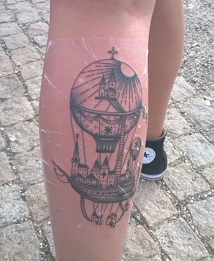 Tattoo of a whimsical architectural scene inside a hot air balloon on a person's calf.
